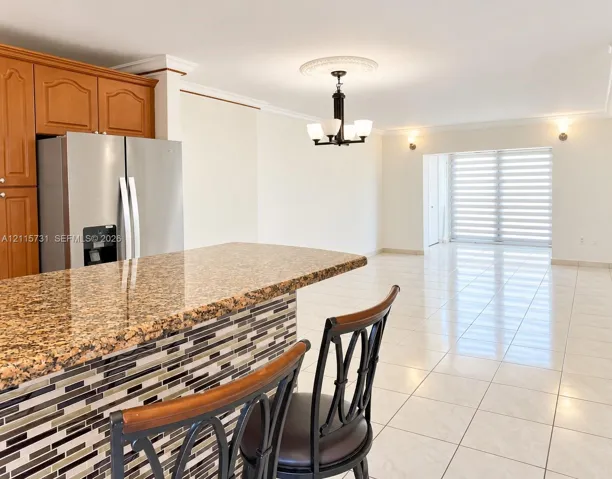 Kitchen island featuring stone countertop and decorative multi-toned tile base, wood-finish cabinetry, stainless steel refrigerator, and tiled flooring