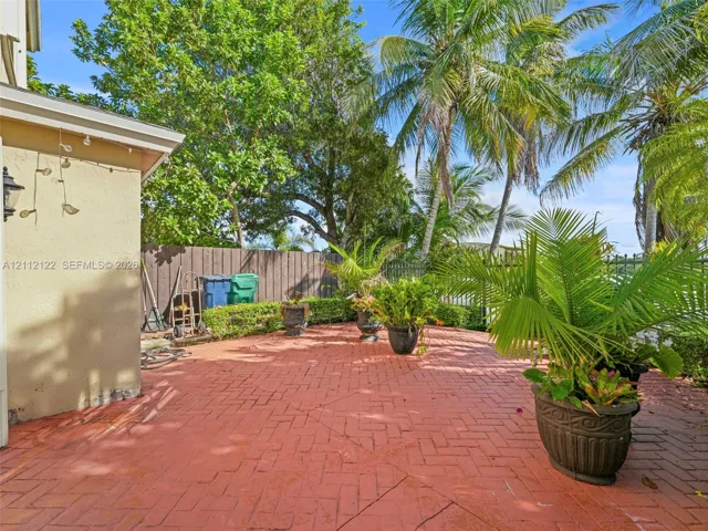 Herringbone-patterned brick patio featuring a perimeter hedge, mature palm trees, and metal fencing
