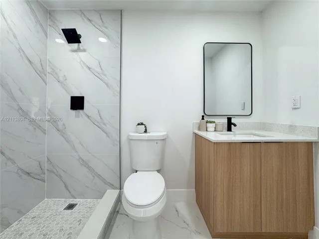 Full-height shower surround with veined stone-finish tile, pebble-tile shower floor, matte black shower fixture, light wood-finish vanity with stone-finish countertop, and wall-mounted rectangular mirror with a black frame