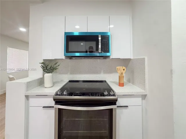Gloss-finish white cabinetry featuring flat-panel doors, stone-composite countertops, high-back matching backsplash, stainless steel electric range, and over-the-range microwave