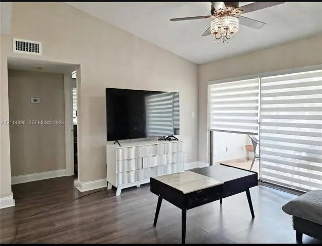 Vaulted ceiling featuring a central ceiling fan with integrated crystal chandelier, wood-finish flooring, and sliding glass doors with roller shades
