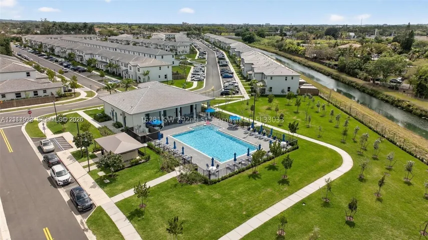 Community swimming pool with a surrounding paved deck, covered pavilion structure, manicured green space, and concrete walking paths