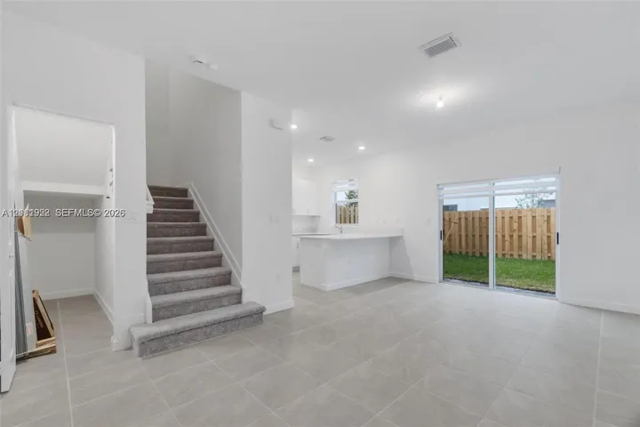 Open-concept interior featuring large-format neutral tile flooring, carpeted staircase, kitchen with white cabinetry and breakfast bar, and sliding glass doors with access to a fenced rear yard