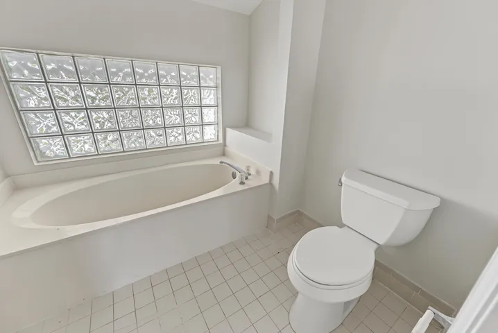 Oval soaking tub with integrated deck, glass block window, chrome plumbing fixtures, and ceramic tile flooring