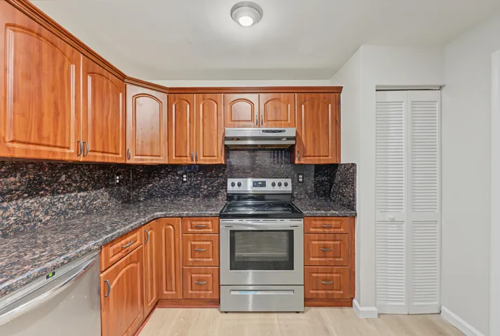 Kitchen featuring wood cabinetry, stone-surface countertops, stone backsplash, stainless steel range with overhead hood, and light-tone wood-finish flooring