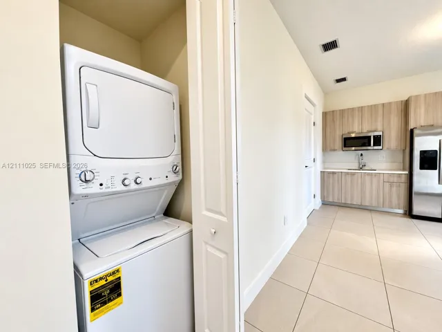 Stacked laundry unit concealed by bi-fold doors, wood-finish cabinetry with stainless steel appliances, and large-format tile flooring