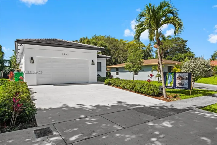 Single-story exterior featuring white stucco siding, a two-car garage with a sectional door, outdoor wall-mounted fixtures, and a concrete driveway surrounded by professional landscaping
