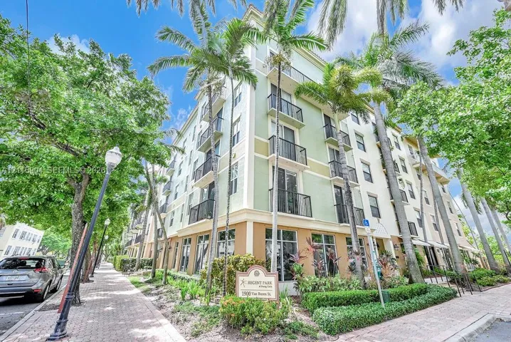 Multi-story building exterior featuring a mixed-tone facade, black metal balcony railings, floor-to-ceiling glass windows, and mature palm landscaping