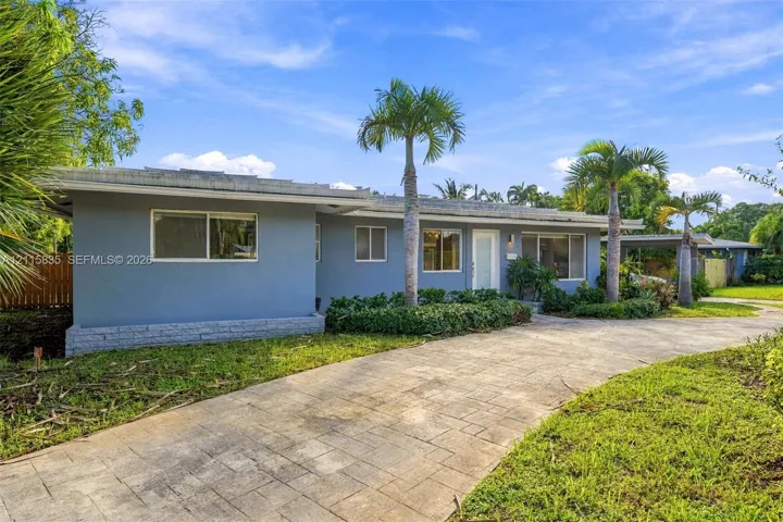 Single-story residence featuring a gray stucco exterior, stone-patterned concrete driveway, mature palm trees, and an attached carport