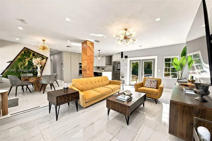 Open-concept living space featuring large-format stone-finish flooring, a decorative geometric wood-paneled structural column, recessed ceiling lighting, multi-arm metallic light fixtures, and glass-paned double doors
