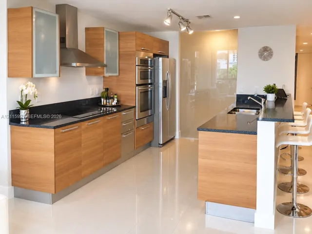 Modern kitchen featuring wood-finish cabinetry, stone countertops, stainless steel appliances, a built-in wall oven, and a breakfast bar with an integrated sink