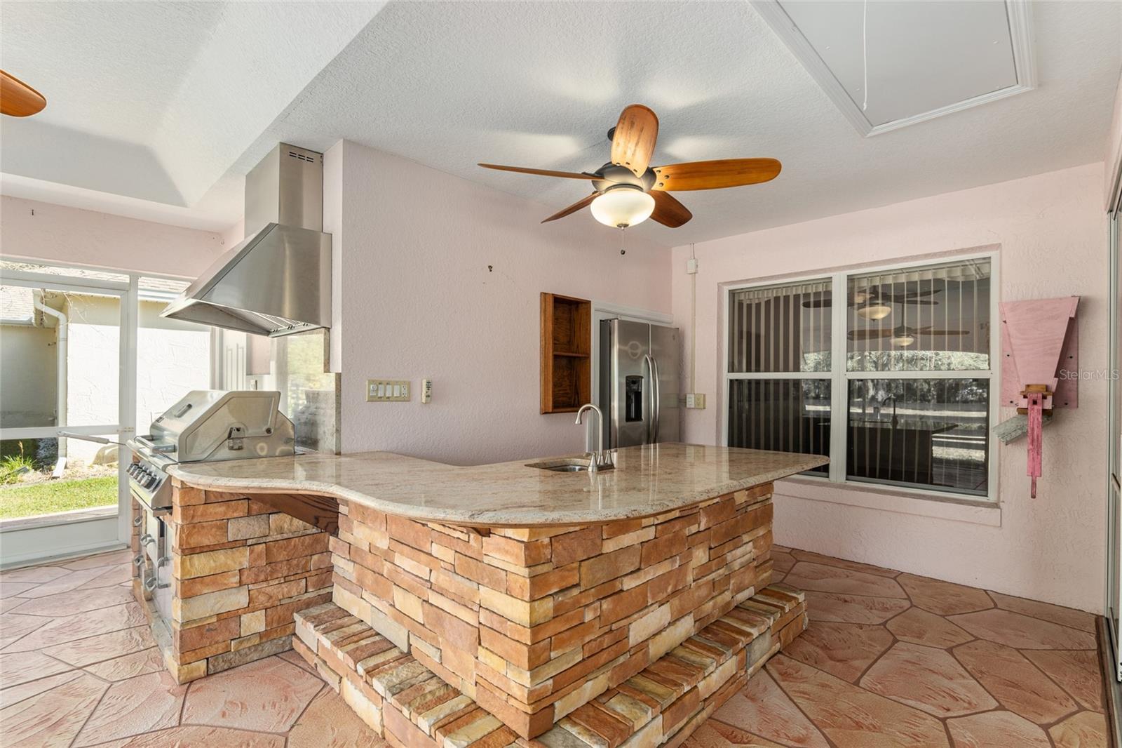 SUMMER KITCHEN in SCREEN ENCLOSED LANAI #2 - Note JENN AIR GRILL w/ exhaust hood, matching CREMA BORDEAUX GRANITE COUNTERTOP, stone base, and side-by-side SS Refrigerator (center back). ATTIC PULL DOWN STAIRS #2 in upper R corner of photo.