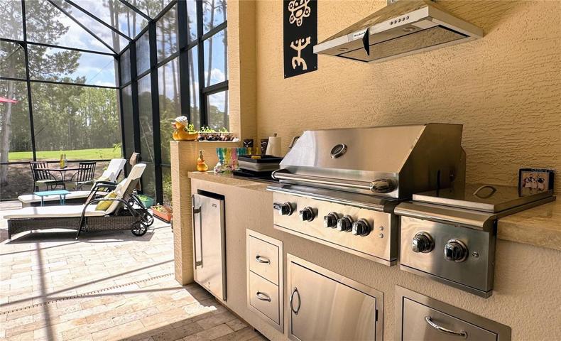 Indulge in the epitome of outdoor culinary convenience – a captivating photo showcasing the remarkable features of your summer kitchen.  The stainless steel LP gas grill and cooktop take center stage, promising sizzling delights with each flame-kissed creation. A vented hood stands as a guardian against smoke, ensuring a comfortable and enjoyable cooking experience. The travertine countertop exudes elegance while providing a spacious surface for prep and presentation. A refrigerator beckons with refreshments, while storage drawers neatly house your culinary essentials. With every element thoughtfully designed, your summer kitchen becomes a haven for both aspiring chefs and those who appreciate fine al fresco dining.
