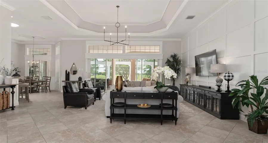 View of LIVING ROOM from FOYER shows octagonal TRAY CEILING w/ CROWN MOLDING, custom WOOD WALL PANELING (R), specialty woodworking trim around TRANSOM WINDOW, & WOOD CORNICE over stack back SLIDING GLASS DOORS leading to massive OUTDOOR LIVING AREA.