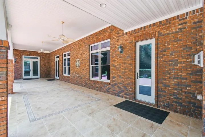 Spacious lanai partially covered by a roof, featuring two ceiling fans and built-in lighting. The entire covered area showcases custom tile flooring, creating an ideal space for entertaining. Access is provided through three French doors and one additional door connecting to various rooms in the home.