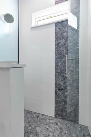 The walk-in shower features a striking pebble-style tile design with a vertical accent strip and coordinating flooring, complemented by a frameless glass door and transom window for natural light.