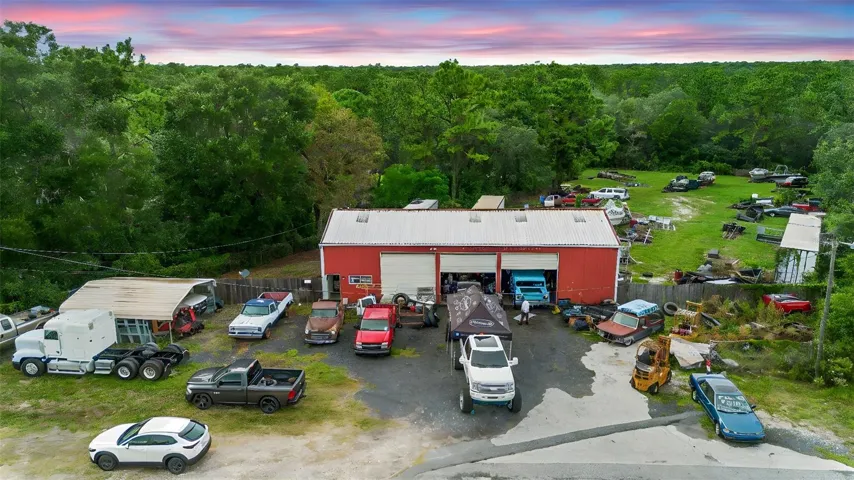 Spacious red commercial shop with multiple bays, ample parking, and storage area, situated on a large lot with mature trees in the background.