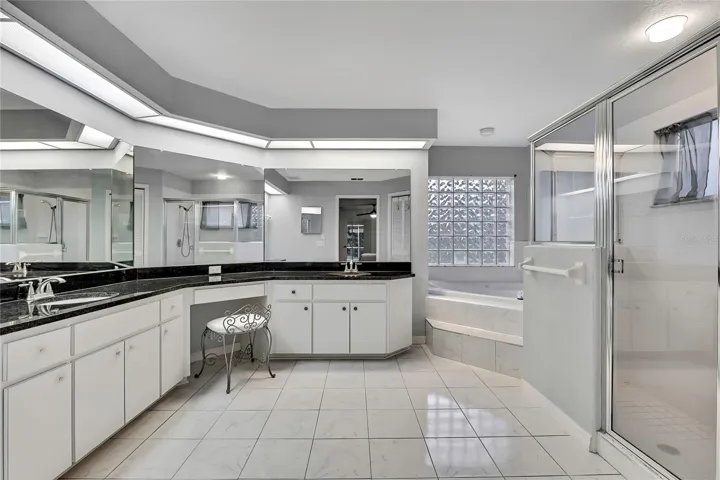 Primary bathroom has long bright double sink granite countertop vanity with vanity seating area.  Glass block wall behind the jetted garden tub provides stylish lighting and privacy.  Large glass enclosed walk in shower.  Luxurious pretty marble look tile flooring accents the white cabinets.