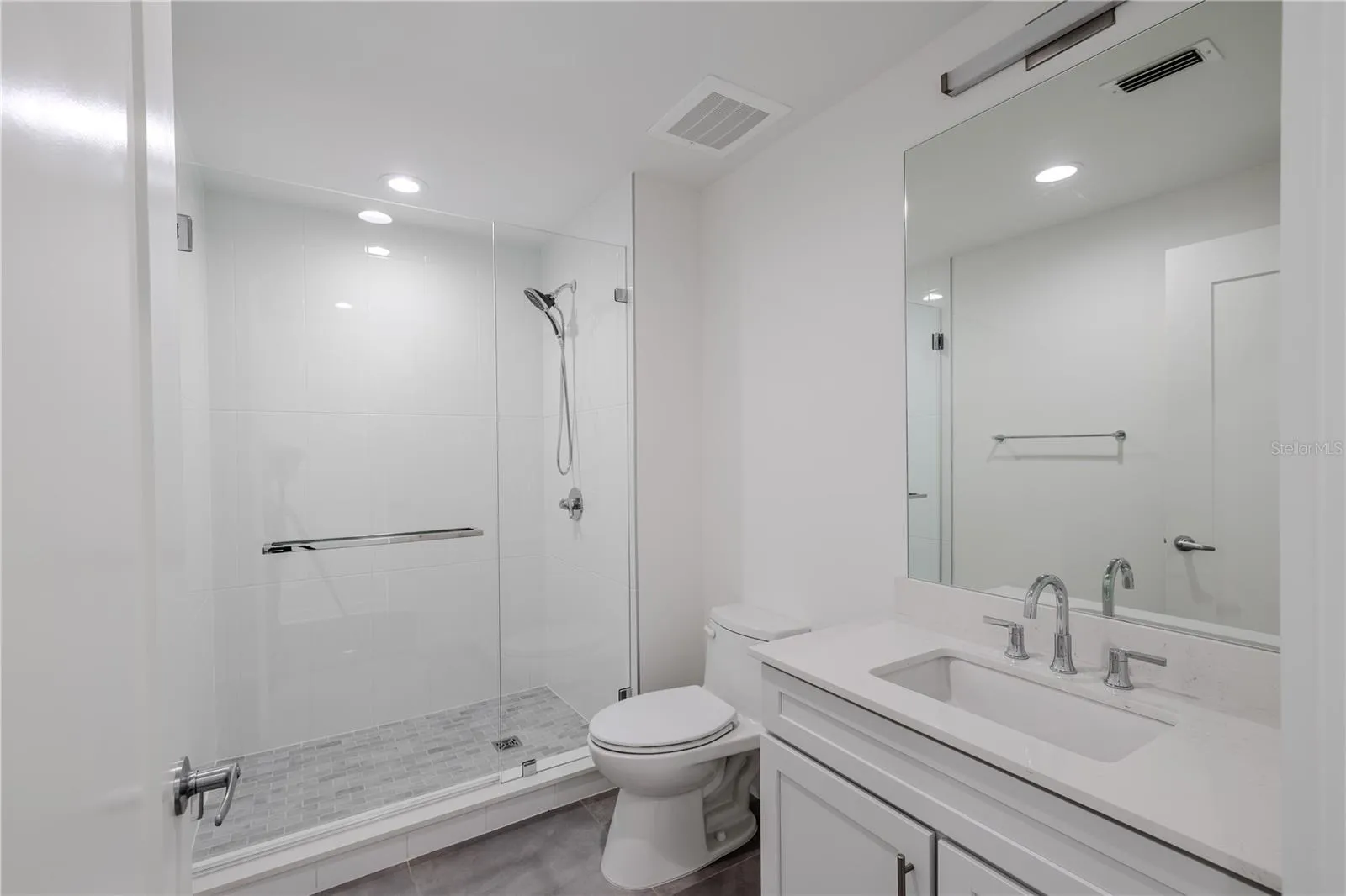 Bathroom 2 offers a sleek, contemporary design with a frameless glass-enclosed walk-in shower featuring large-format white tile and a mosaic tile shower floor. A quartz-topped vanity with under-mount sink, polished chrome fixtures, and a wide mirror with integrated lighting create a bright, spa-like atmosphere. Clean lines and neutral tones make this en-suite both elegant and functional—perfect for guests or as a private bath for the secondary bedroom.