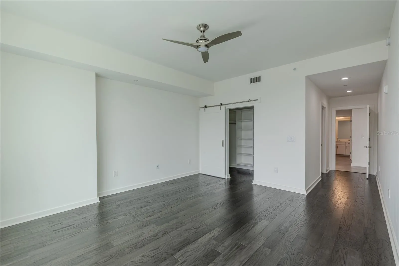 This inviting second bedroom features rich hardwood floors, recessed lighting, and a sleek ceiling fan that adds a touch of modern style. A sliding barn door opens to a custom closet with built-in shelving for organized storage. The room connects conveniently to an en-suite bath