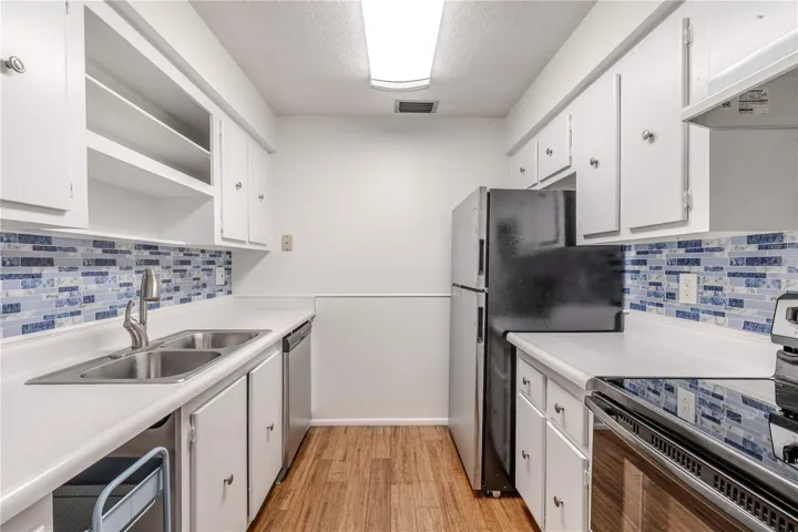 The kitchen features a clean, functional layout with crisp white cabinetry, stainless steel appliances, and a double sink for added convenience. The blue mosaic-style backsplash adds a fresh coastal touch and beautifully complements the warm bamboo flooring. Ample counter space and open shelving provide both practicality and charm, making meal prep and storage easy.
