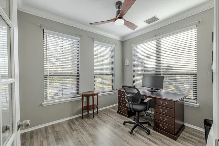 Bright flex room/home office with crown molding, modern ceiling fan, and wraparound windows that fill the space with natural light, complemented by updated flooring.