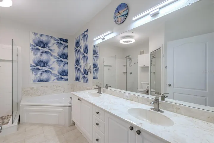 This view highlights the spa-inspired primary bathroom with a glimpse of the glass-enclosed walk-in shower featuring pebble-style flooring, a deep soaking tub with elegant tile surround, and bright, contemporary finishes throughout.