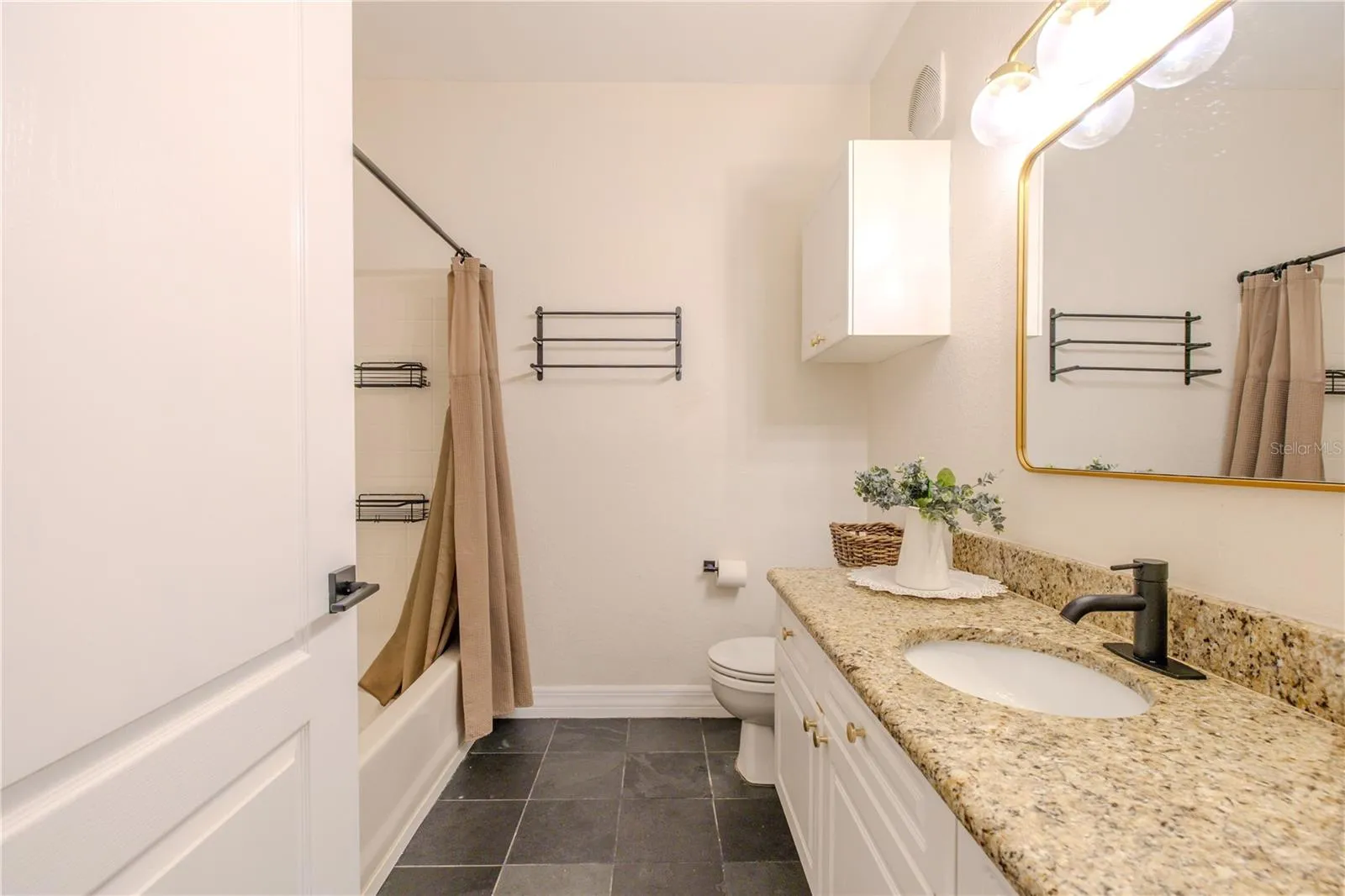 Another view of the clean, bright, and thoughtfully updated, featuring a granite-topped vanity with an under mount sink, crisp white cabinetry, and a matte black faucet for a modern contrast. A framed mirror and contemporary vanity lighting add warmth and style, while the tub/shower combination offers classic tile surround and functional shelving. Dark tile flooring grounds the space, and neutral finishes throughout create a fresh, timeless look that’s both practical and inviting.