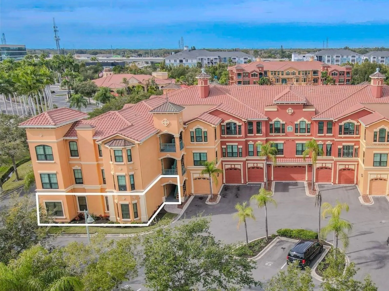 This aerial view highlights the striking Mediterranean-style architecture of Grand Bellagio at Baywatch, showcasing its warm stucco façade, decorative architectural accents, and classic barrel tile roofing. The building’s symmetrical design is anchored by elegant towers, arched openings, and covered breezeways that create a cohesive, resort-inspired aesthetic.