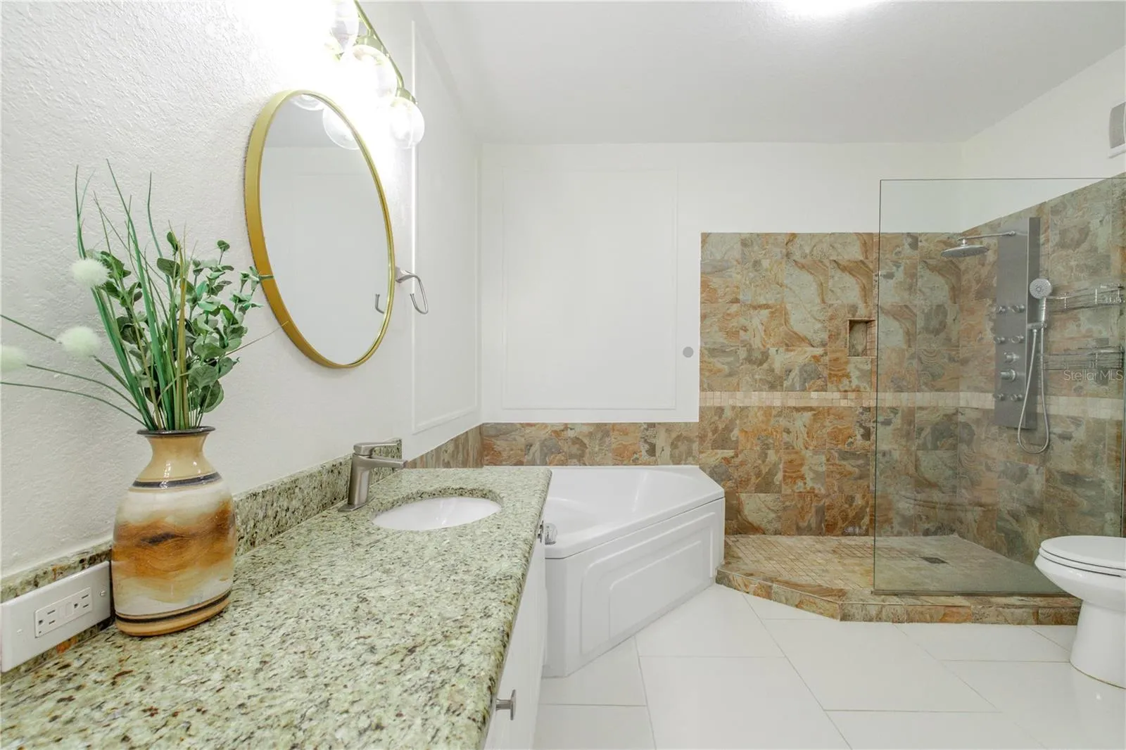 The primary ensuite bathroom is designed with a spa-inspired layout that includes a granite-topped vanity with undermount sink, crispcabinetry, and elegant lighting paired with a framed mirror. A soaking tub anchors the space, while the walk-in glass-enclosed shower features natural stone tile, a built-in niche, and a multi-function shower system for a true retreat experience.