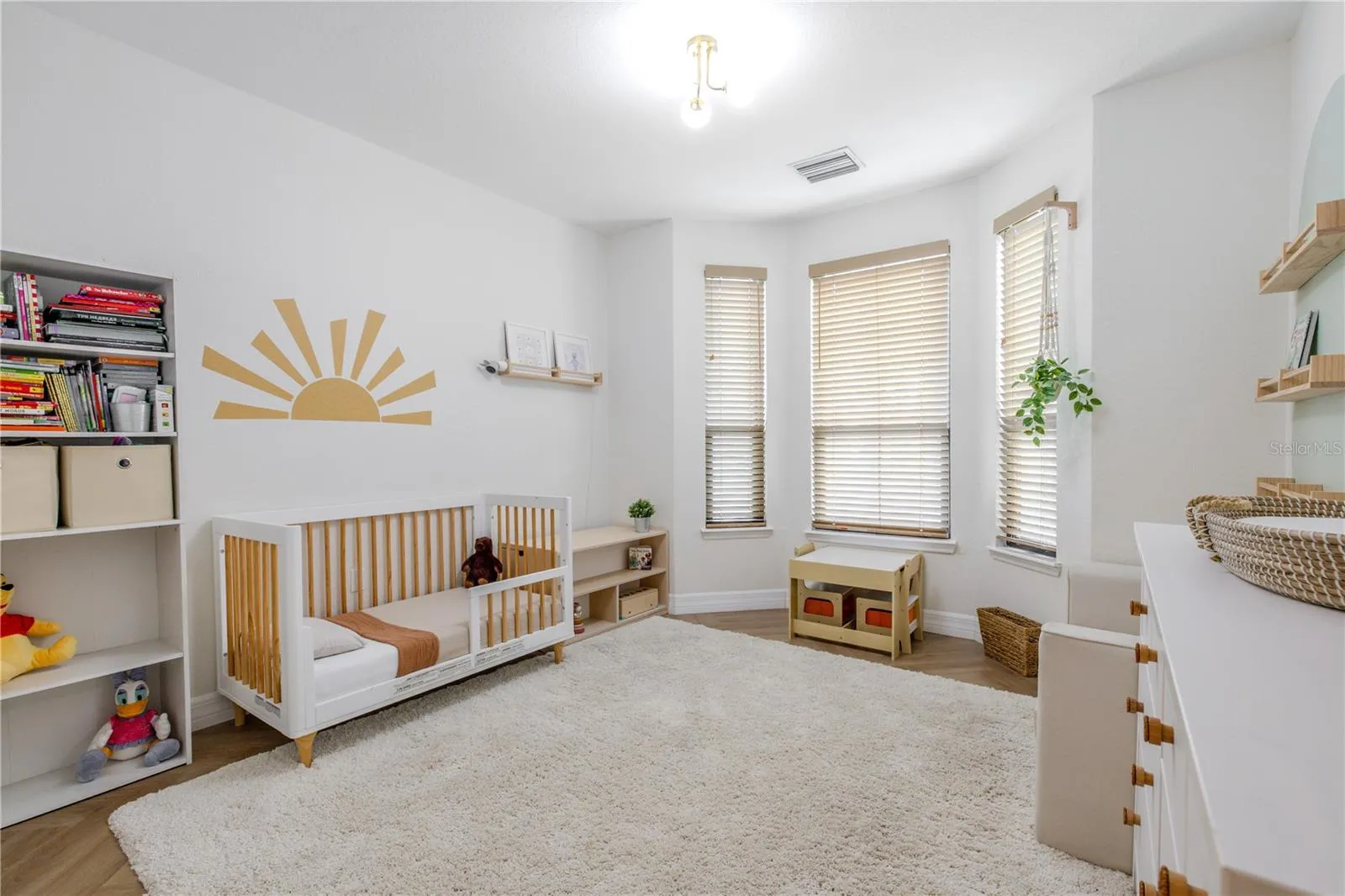 Another view of the second bedroom is bright, airy, and thoughtfully styled, featuring soft neutral walls, warm wood-look luxury vinyl flooring, and abundant natural light from multiple windows fitted with classic wood blinds. The spacious layout easily accommodates a nursery, guest room, or home office, with room to grow and reconfigure over time.
