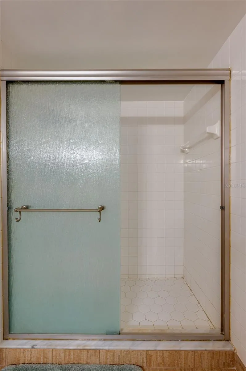 Close-up view of the primary suite’s shower showcases a hexagon tile floor and full tile surround behind frosted sliding doors.