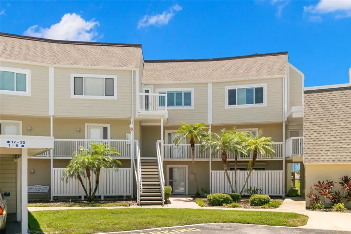 Well-kept coastal-style townhome building with light neutral siding, covered walkways, and a landscaped courtyard entry framed by mature palms. Front door sidelights have been installed with second thickness of glass for improved electric conservation with increased insulation compared to the original single glass thickness.