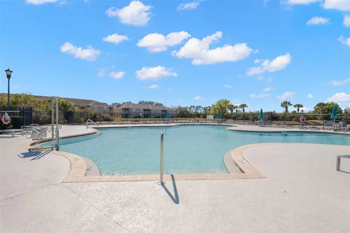 Resort-style community pool featuring a spacious deck with ample lounge seating and shaded areas. The inviting, freeform design offers both shallow lounging space and deeper swim areas, perfect for relaxing or enjoying Florida sunshine year-round. Securely fenced with surrounding greenery and open skies, creating a private yet welcoming atmosphere for residents.