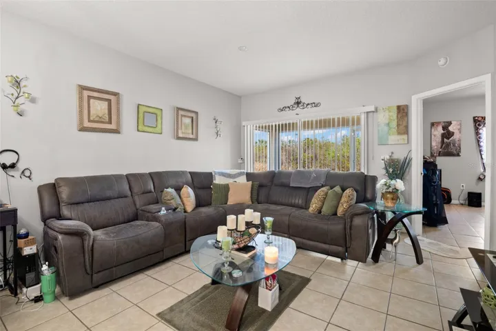Generously sized living room with ceramic tile flooring and large sliding glass doors that fill the space with natural light while offering peaceful greenery views. The layout easily accommodates full-size furnishings and maintains an open, airy feel. Sliding doors lead directly to the screened lanai, enhancing indoor-outdoor living.