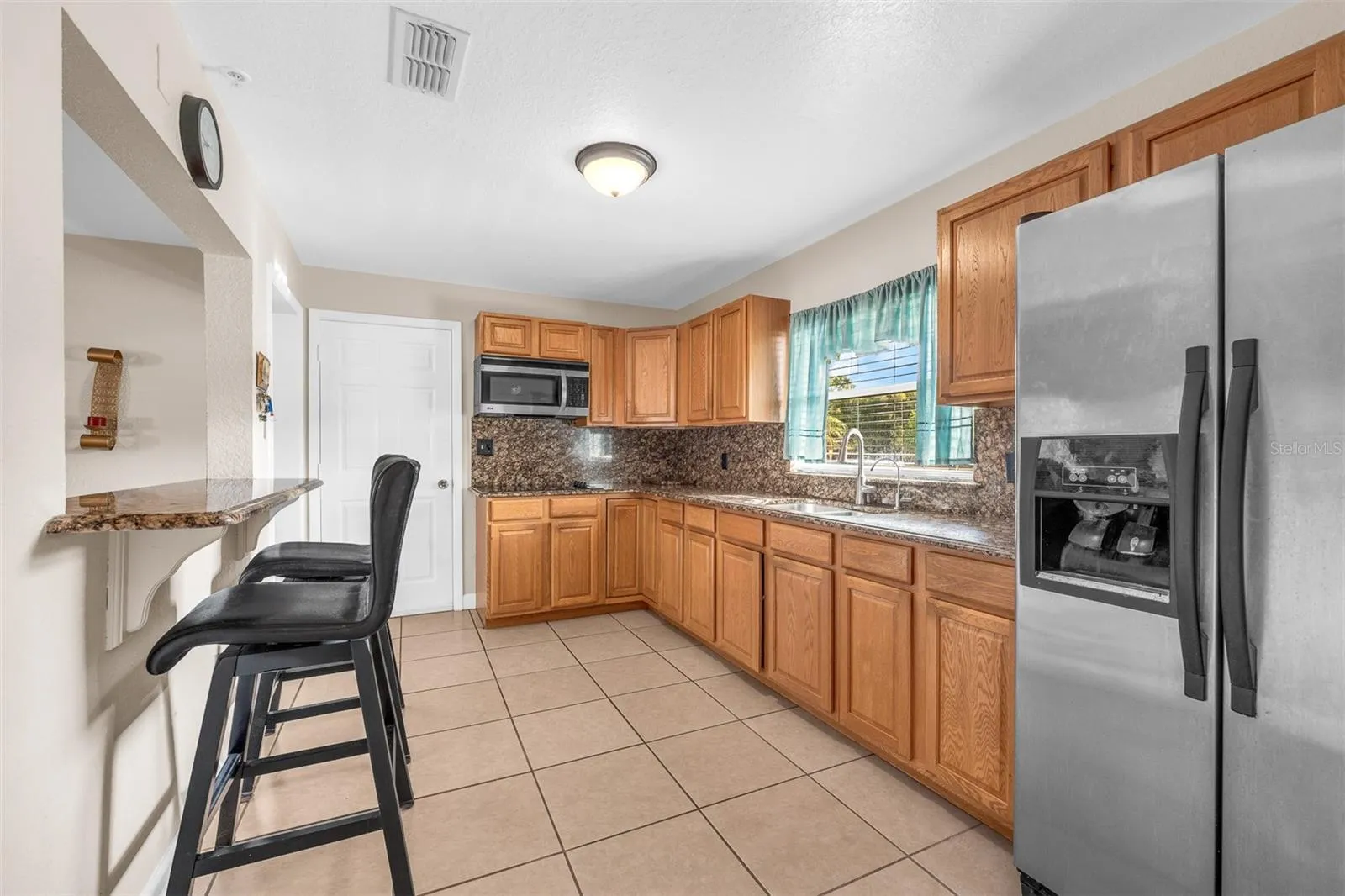Another view of the kitchen showing the cooktop, built-in microwave, granite backsplash, and side-by-side refrigerator. Natural light from the window brightens the workspace.