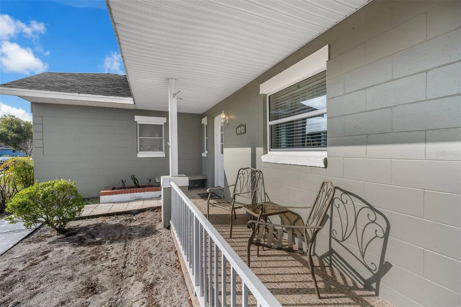 Tiled front porch with white railing, two seating chairs, and a glass-panel entry door. Includes exterior lighting and front-facing windows.