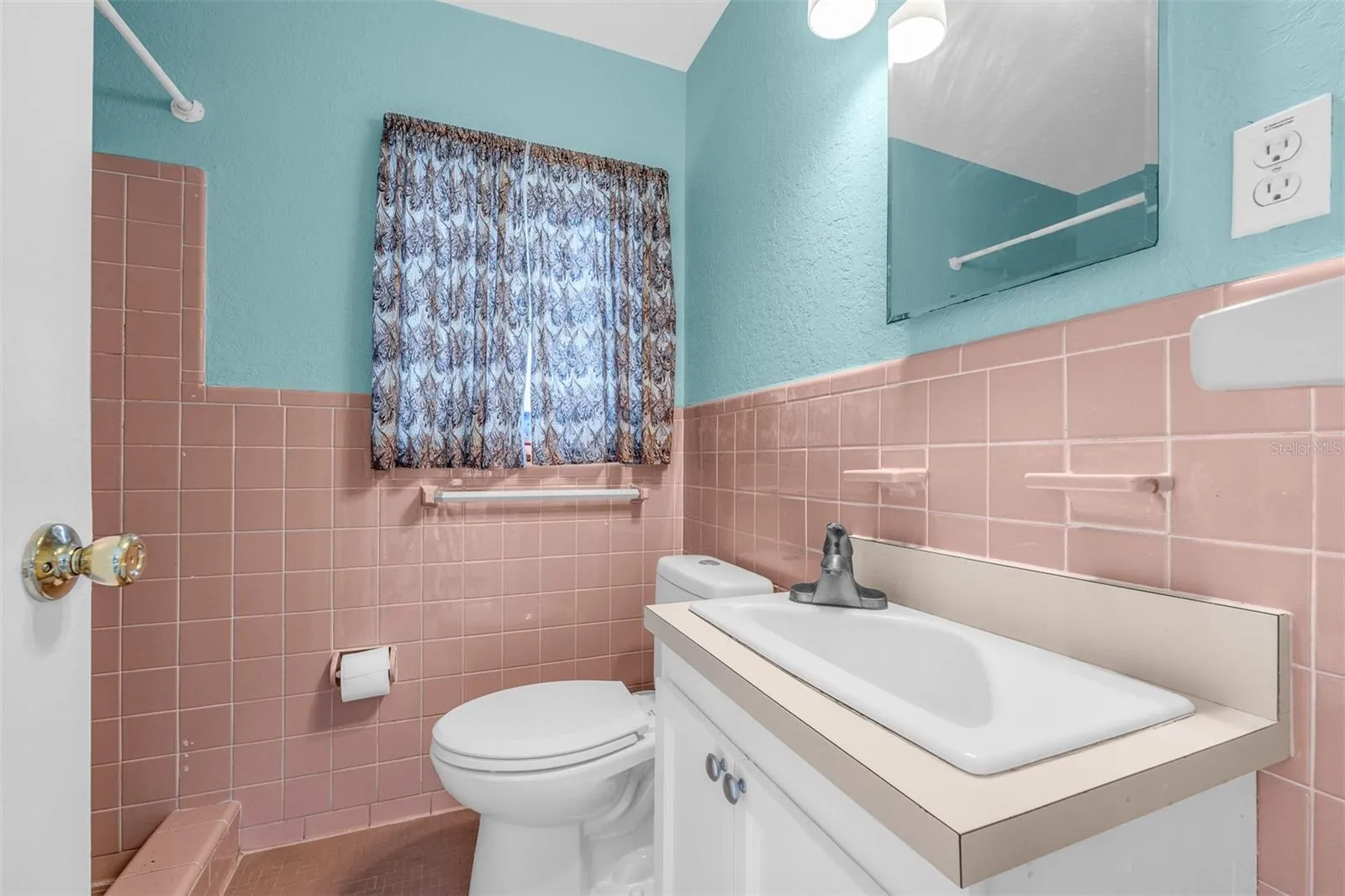 Bathroom with retro tile accents, white vanity with storage, overhead lighting, and a window with privacy curtain. Features a combination of painted upper walls and tiled lower walls with built-in ceramic fixtures.