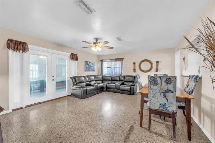 Open living and dining area with polished flooring, large sectional seating, and double doors leading to an additional flex space. Neutral wall colors and multiple windows provide natural light.