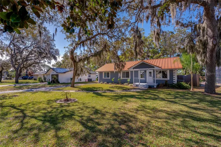 Wide front exterior view showcasing the expansive front lawn, mature trees, and attractive metal roof. The spacious setting creates a welcoming first impression.