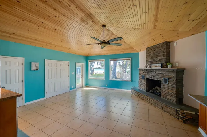 Expansive family room with a vaulted wood ceiling, tile floors, and a striking stone fireplace as the focal point. Large windows frame the backyard and water views beyond.