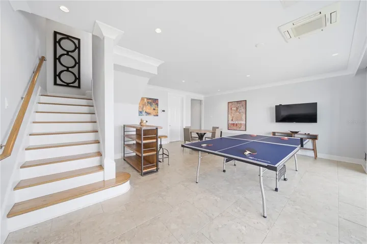 The lower-level includes a two-car garage and a spacious foyer/media/game room featuring a mounted TV and floor-to-ceiling multi-panel sliding glass doors that create a dramatic wall of glass, instantly framing tranquil water views.