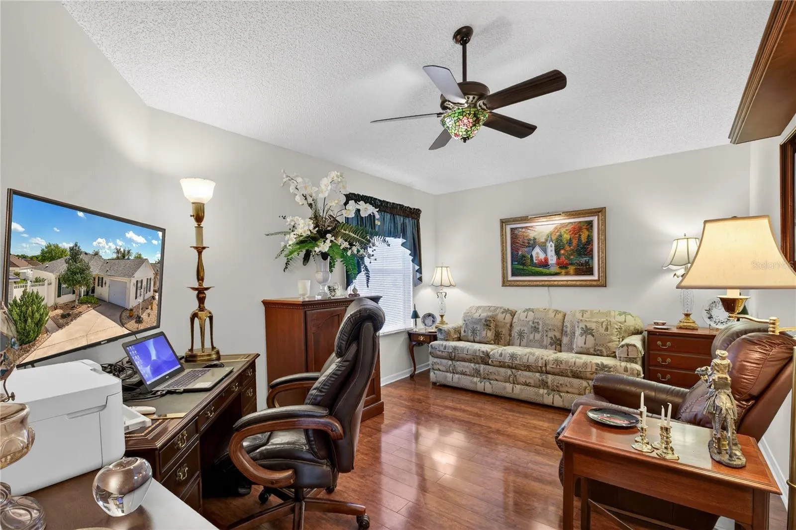 This expansive second bedroom offers a comfortable and inviting space, highlighted by gleaming laminate wood floors. The room is further enhanced by a stylish ceiling fan, complete with a beautiful Tiffany glass light shade. An electric fireplace adds a touch of cozy elegance,.