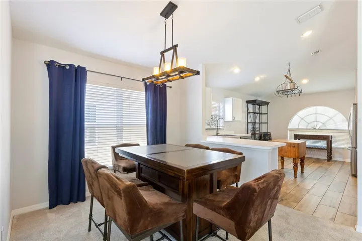 Bright open-concept dining area with stylish chandelier, large window with blue curtains, and seamless flow into a modern kitchen featuring a breakfast bar and abundant natural light.