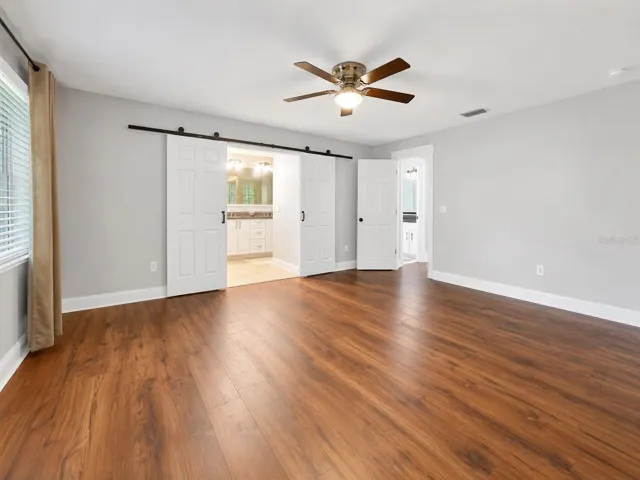 Spacious primary suite with warm wood-like flooring, soft natural light, and charming barn doors leading into the private bathroom retreat.