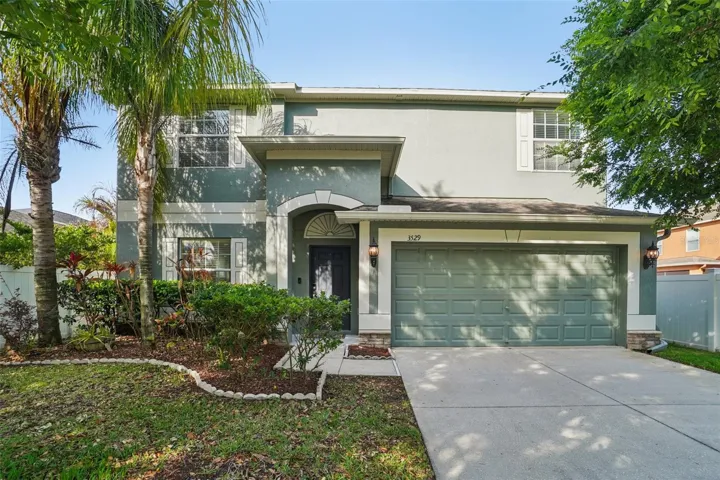 Well-maintained two-story home with sage green stucco exterior, arched entry detail, two-car garage, and lush tropical landscaping with mature palms.