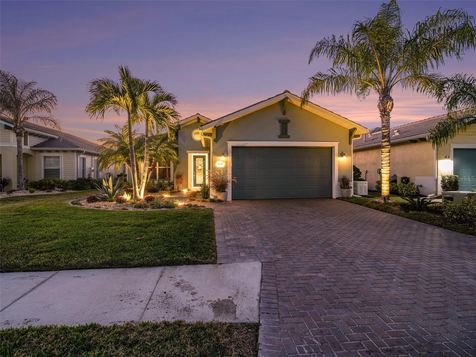 Twilight front exterior with tropical landscaping, paver driveway, and warm lighting that enhances curb appeal.