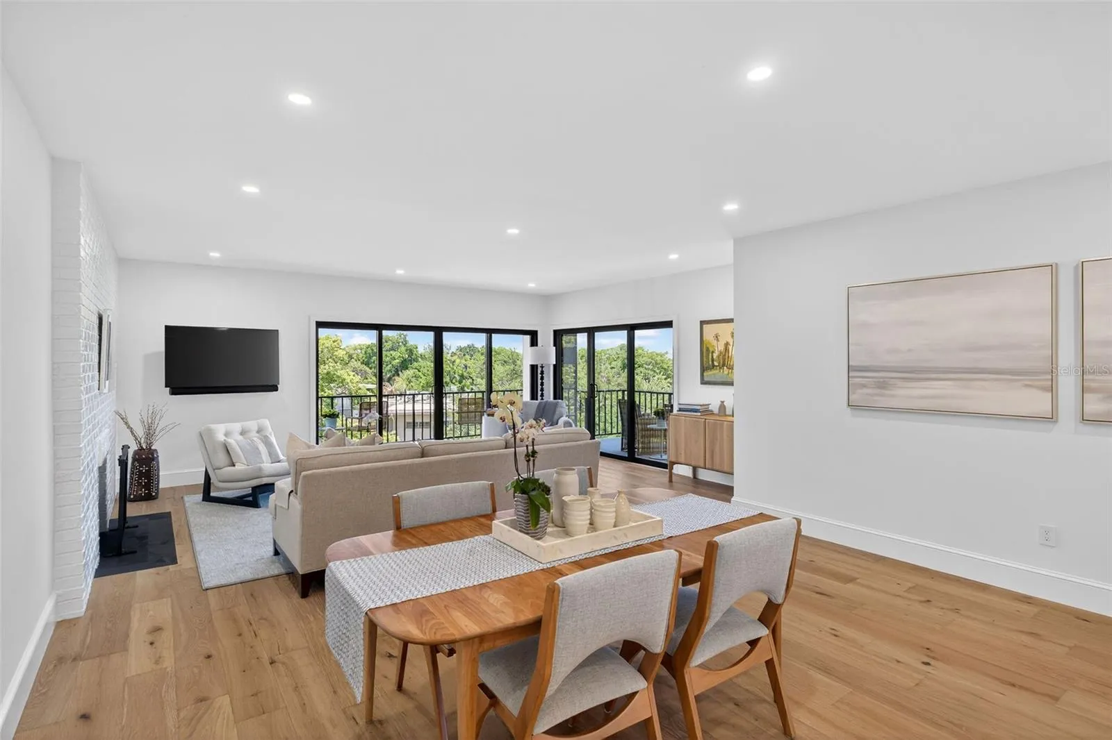 Dining space unfolds toward walls of glass and a private balcony, where warm wood floors, a sleek fireplace, and treetop views create the perfect backdrop for anything