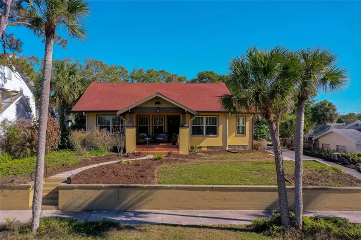 Classic bungalow-style exterior with a welcoming covered porch, red roof, and mature palm trees enhancing curb appeal. Elevated lot with a defined walkway adds character and presence.
