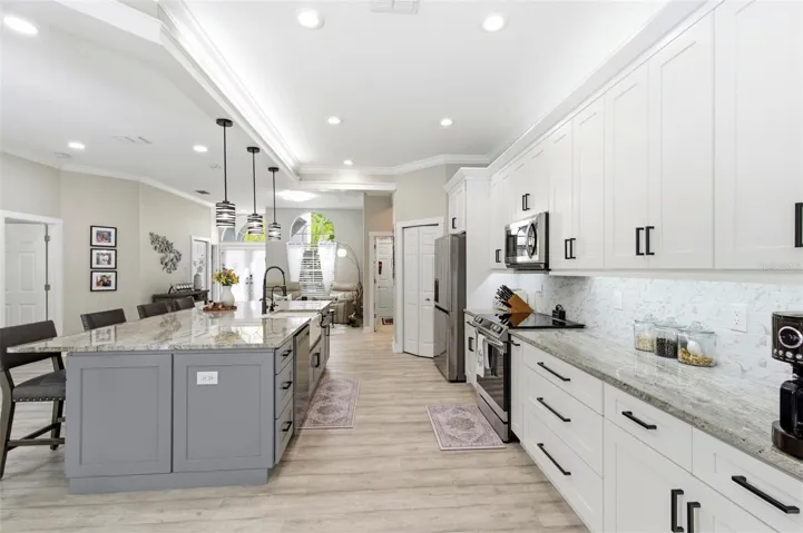 Beautifully designed open-concept living space showcasing a spacious chef’s kitchen with a large granite island, ample seating, and abundant cabinetry. Stainless steel appliances, modern pendant lighting, and elegant finishes create a clean, upscale feel. The layout flows seamlessly into the living and dining areas, with sliding glass doors opening to the pool area—perfect for indoor-outdoor entertaining and everyday living.